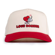 A white baseball cap with a red brim featuring a graphic design of a heart and bomb with the text 'LOVE BOMBER' on the front.